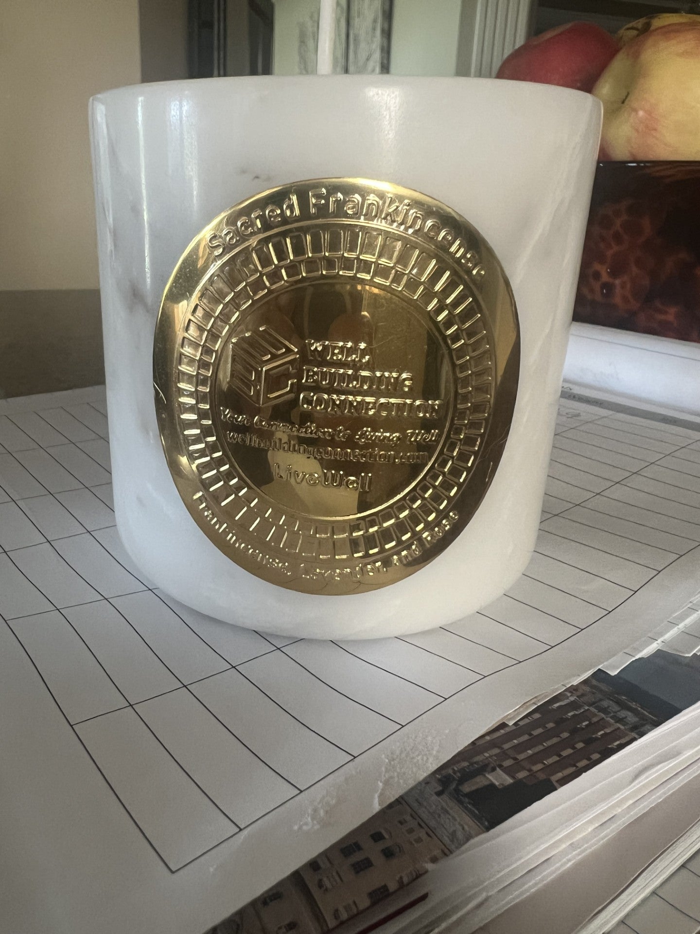 A white marble candle jar with a large circular gold-toned medallion on the front, placed on a lined sheet of paper with a blurred indoor background.