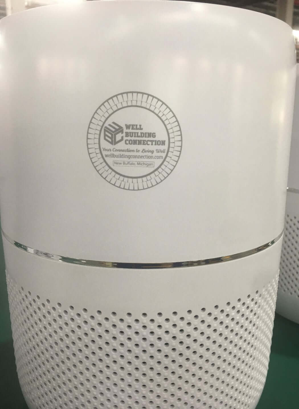 A white cylindrical HEPA air purifier with a perforated lower section and a Well Building Connection logo on the front. Indoor Air Pollutants too many to list and everyone benefits from this air purifier from Well Building Connection.